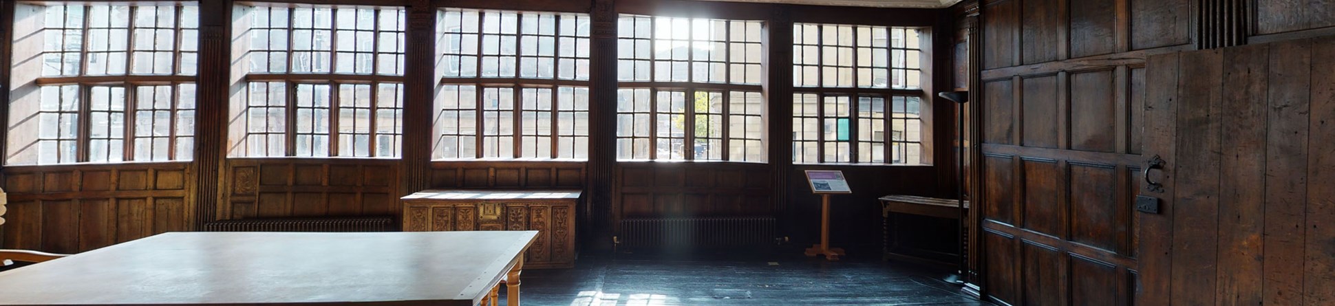 A historic interior with wooden panelling and a stuccoed ceiling, illuminated by a range of historic windows.