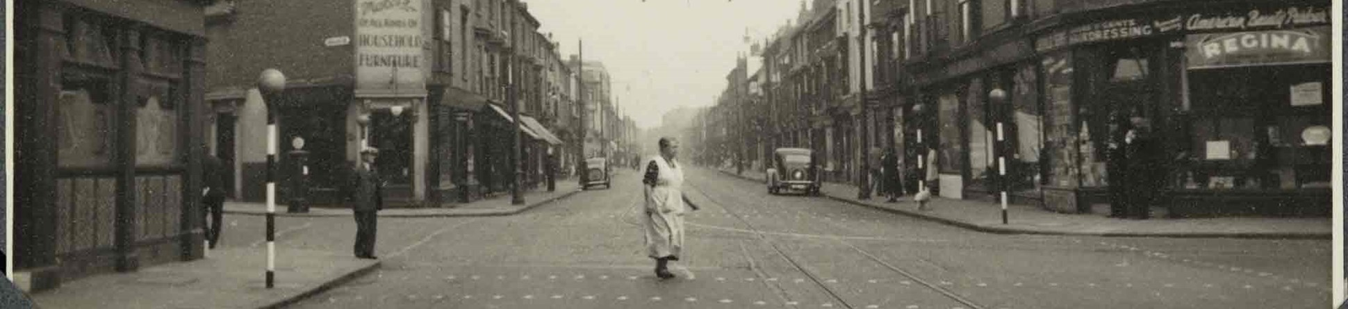 Black and white photograph of an urban street scene featuring a woman in an apron using a pedestrian crossing. Three-storey buildings flank the road, most of which have ground floor shops. The upper storeys of some buildings show painted signs and advertisement boards. A label applied to the bottom-right of the photograph reads: 'Birmingham. Sherlock Street', part of which is obscured by one of four corner photograph album mounts. 