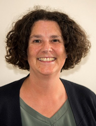 Photograph in portrait style, showing dark, curly haired woman smiling widely wearing a green top and black cardigan. 