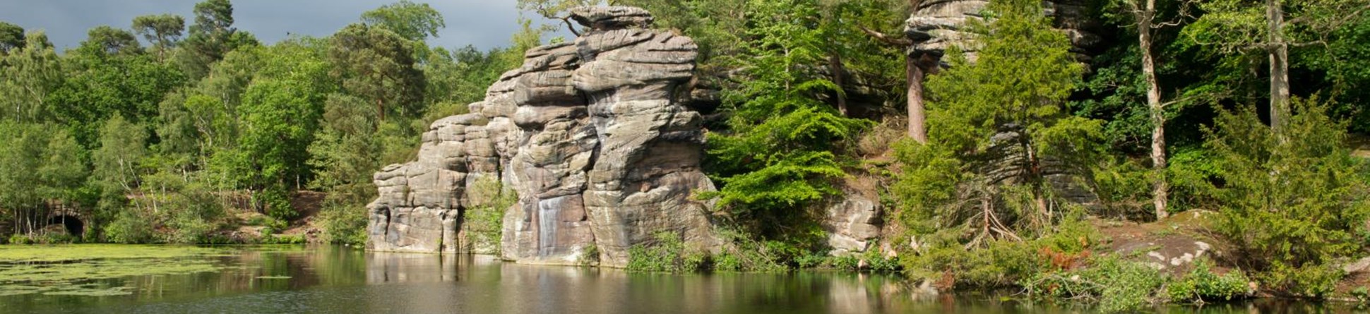 Lake with water lillies in the foreground bordered by dramatic rock features and trees.