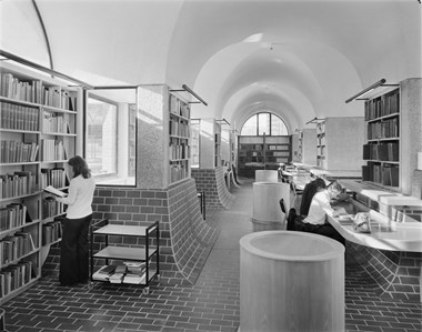 A black and white photo showing the interior of the Guildhall School of Music and Drama Library. The library’s floors and walls are partially covered with bricks, and three people are reading books in the area.