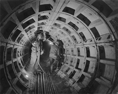 A black and white photo showing two workmen in a large tunnel at the Barbican site. The men are working on reinforcements with a few lights installed in the tunnel.