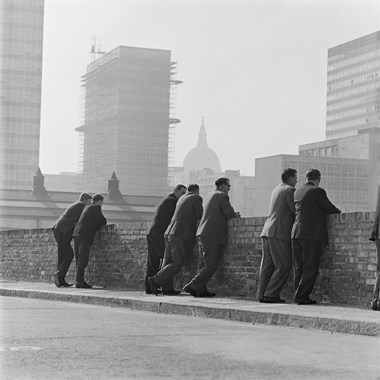 A black and white photo showing a group of men in suits leaning against a brick wall. The background shows the dome of St Paul’s Cathedral, as well as office blocks.