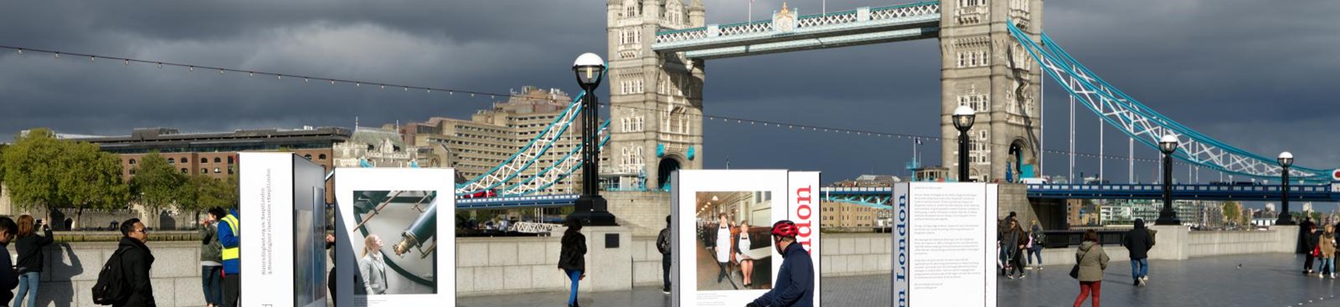 Tower Bridge, London with pedestrians and cyclists in the foreground stopping to look at Historic England's I am London exhibition panels in a pedestrianised area on the south bank.