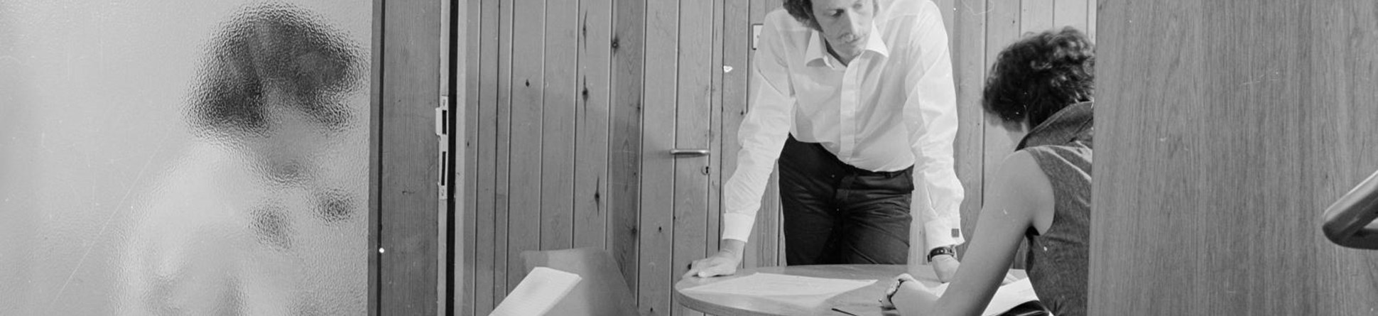 1975 office scene - black and white photo of three people around a meeting table in a timber clad room in the offices of the architects austin smith lord - as seen through a doorway.