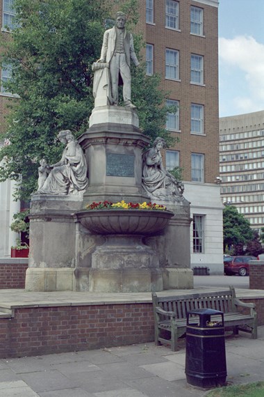 Stone figure of a man in Victorian dress, standing with his hand resting on a Bible. Below, to either side, a seated female figure. To left, Charity holds one infant and comforts another; to right, Peace holds an olive branch, a lamb at her feet.