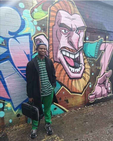 Man wearing headphones and carrying laptop bag standing in front of a bold mural at the Custard Factory.