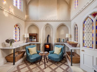 Interior of room with two armchairs and cofee table in the middle. The walls include arched recesses and mullioned windows with stained glass edging.