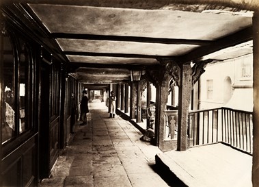 Two men, both wearing bowler hats and one wearing a long apron, stand in one of the covered rows of Chester.