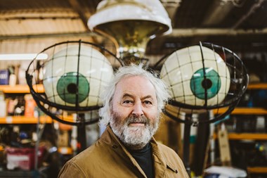 Head and shoulders portrait shot of a man in a brown coat with a shop display of two encaged eyeballs, one either side of his head, behind him.