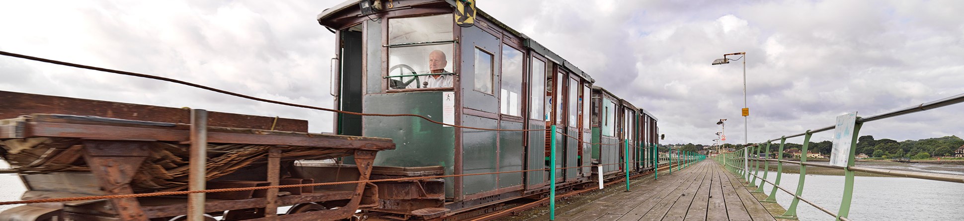 Photo along wooden pier with train on the tracks on the left and iron railing running down the right hand side.