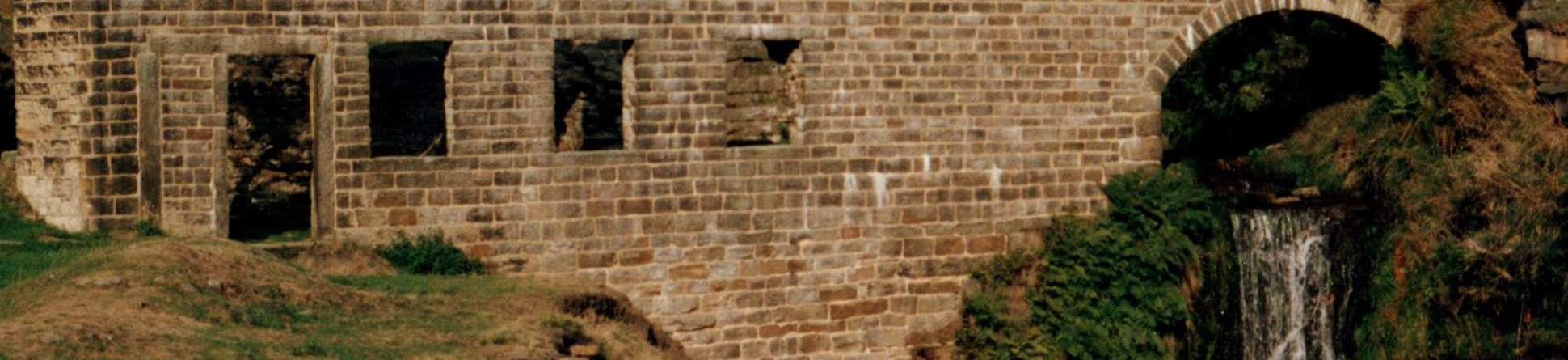 The stone facade of a ruined textile mill, featuring an arch over a waterfall.