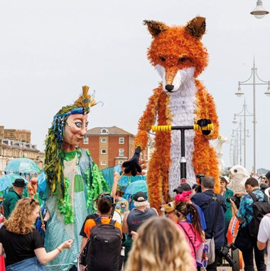 Two large puppets surrounded by people in a street setting.