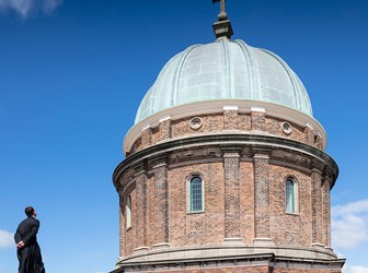 A figure in a priest's cassock stands on a curved roof, looking at the brickwork and copper roof of a domed church.