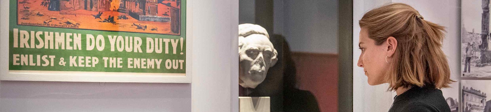 A woman visiting the What Remains exhibition looks into a display cabinet including a sculpture of a man's head that has had the nose knocked off. A poster to her left shows a church in flames and includes the text: "IRISHMEN DO YOUR DUTY! Enlist and keep the enemy out"