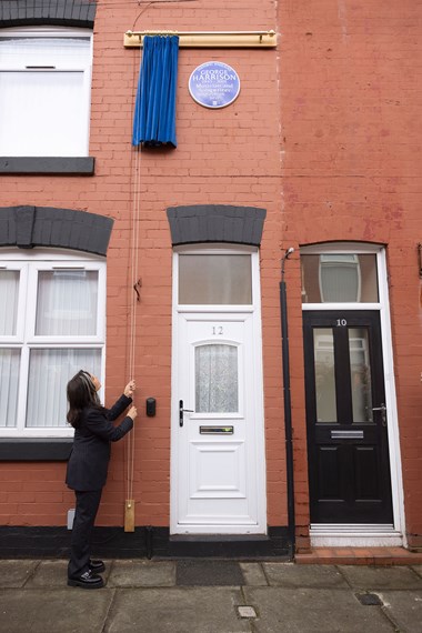 A woman stood in front of a red brick terraced house, pulling a cord to reveal a heritage blue plaque