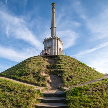 A grassy mound with a tall stone monument rises in front of a blue sky. 