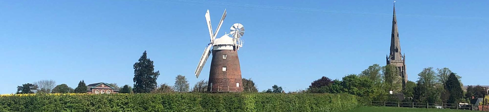 The mill, a red brick building with white sails and top sits in the background behind a field and hedge, adjacent to the village church.