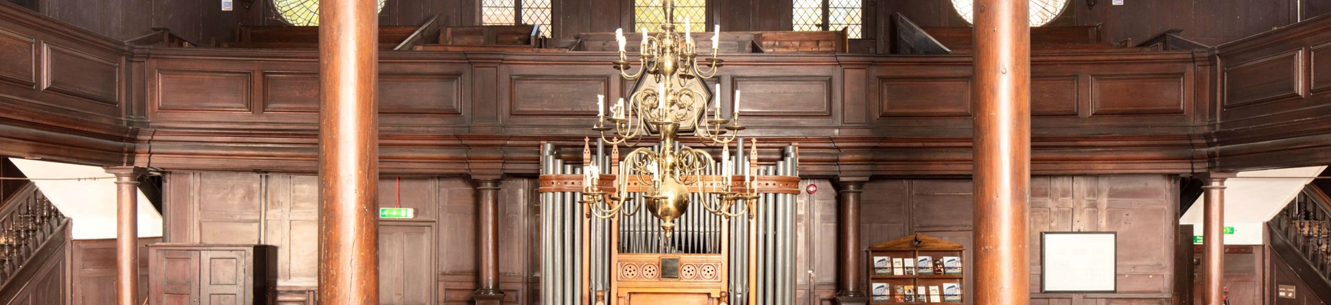 The beautiful interior of the Unitarian Meeting House showing dark wood pews, parquet flooring, wooden pillars and the late 19th century organ. A chandelier hangs from the middle of the ceiling.