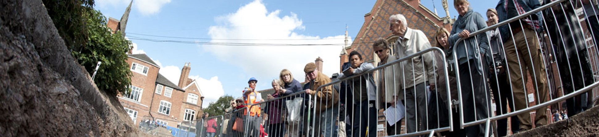 Members of the public peering over fencing into an archaeological trench, photographed from the bottom of the trench looking up.