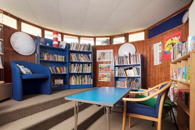 Photo of the interior of a school classroom with bright blue bookshelves and seats. The walls are wood panelled with small windows at the top.