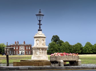 View of a stone drinking fountain with a lamp on top. In front is a stone trough filled with pink flowers.