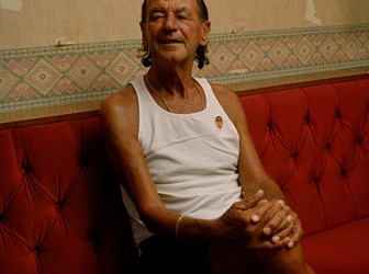 Person wearing a white tank top gold chain bracelet and necklace sits on red booth seating at a men's working club. There is patterned wallpaper behind them.