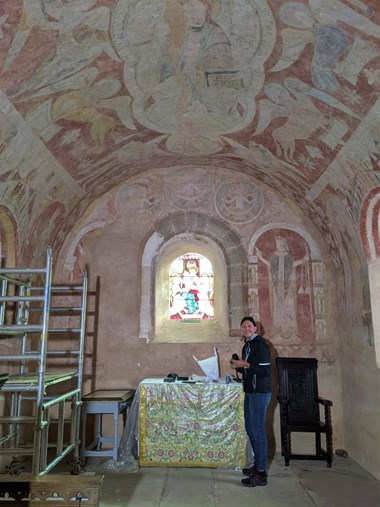 Photograph showing a decorated historic building with arched roof covered in a faded mural. A woman is shown next to equipment, she is dark haired and smiling. 