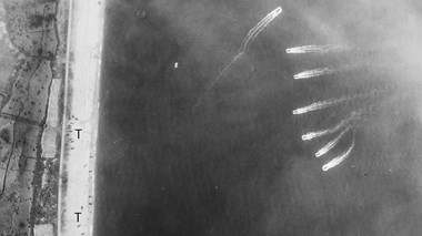 Detail from a black and white vertical aerial photograph, showing a strip of beach bordering a patchwork of fields at the left edge of the image. To the right, six vessels at sea sail in a line towards the beach, trailing wake behind them. Another vessel sails above them in the opposite direction. Vehicles are dotted on the beach. Two letter 'T's applied to the image mark the location of tanks. Wafts of smoke partly obscure the view at the top of the image.