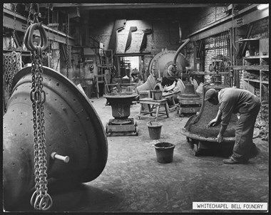 A black and white photograph of the interior of a factory. To the right, a man stoops to work on bell-shaped clay on a wheeled trolley. In the left foreground, chains hang in front of a bell mould.