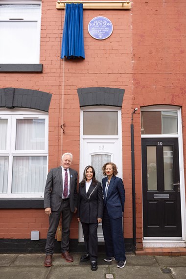 Three people stood on the street outside a red brick terraced house, underneath a heritage blue plaque mounted on the wall at first floor level that has just been revealed from behind a blue curtain