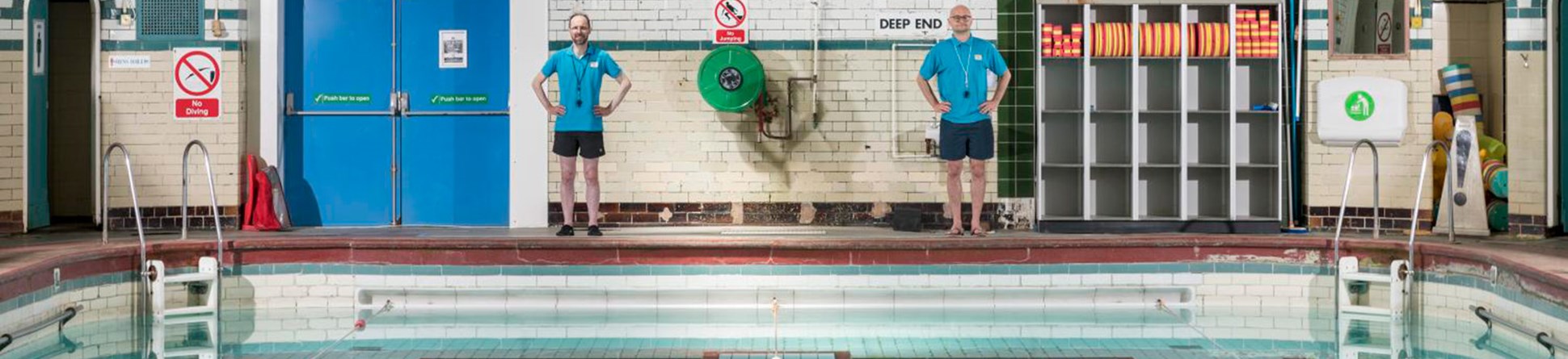 Two lifeguards stand beside an indoor pool marked "deep water," surrounded by tiled walls and large windows.