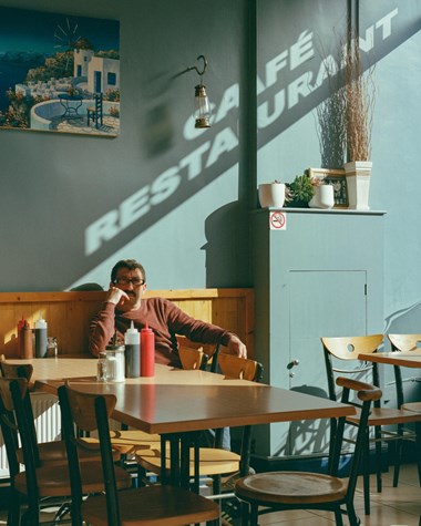 A photograph of a man sitting at a table in a café with blue walls. Above him is a painting of white buildings on a Greek island. The words 'Café restaurant' can be seen above him, the shadow of text from the window (off camera.)