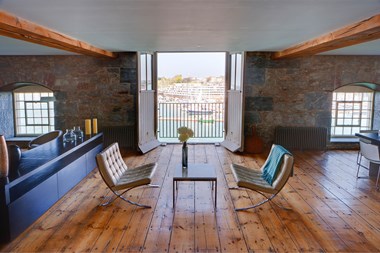 A photograph of an interior living space with a wooden floor, exposed beams and a seating area. Open doors at the back of the room show a view of a harbour.