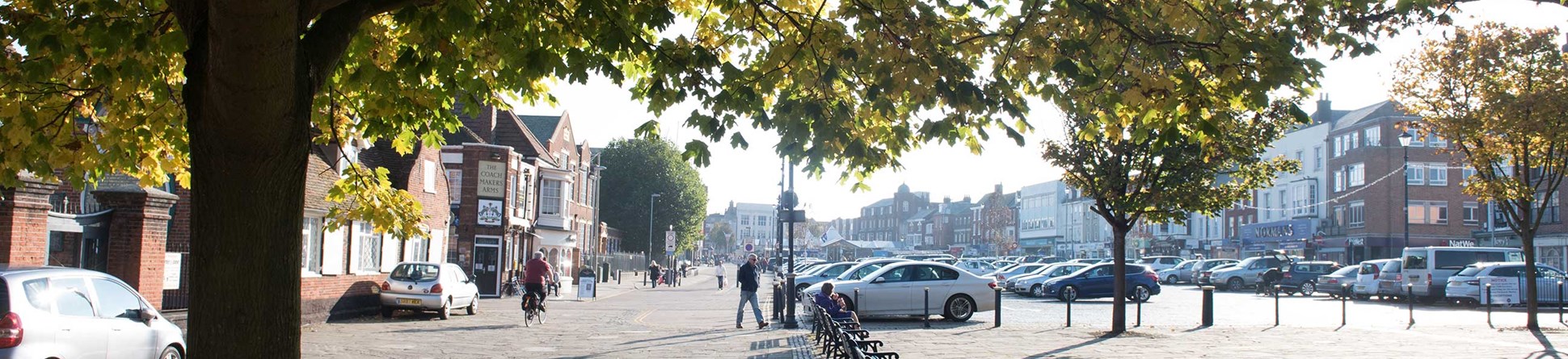 Great Yarmouth Market Place viewed from under a tree. The buildings that surround the market square are in the background. In the middleground are parked cars.