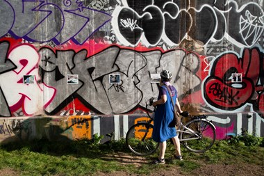 Person standing with bike in front of postcard size photographs mounted on a concrete graffiti wall.