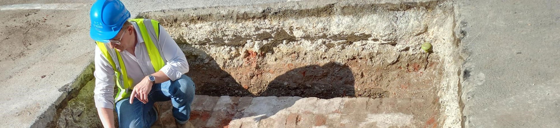 An archaeologist kneeling on a brick arch pointing at earlier masonry within a small archaeological trench.