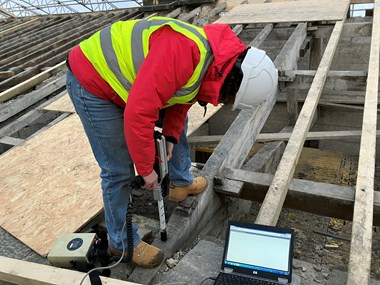 Photograph showing dark-haired woman in red coat, high vis and hard hat using a decay detection drill on Belsay Hall roof trusses. 