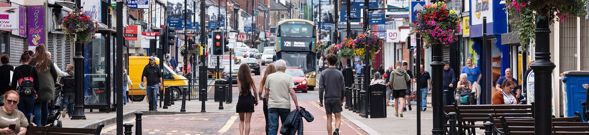 A high street scene featuring people, benches, lamp posts, shops, and traffic in the distance.