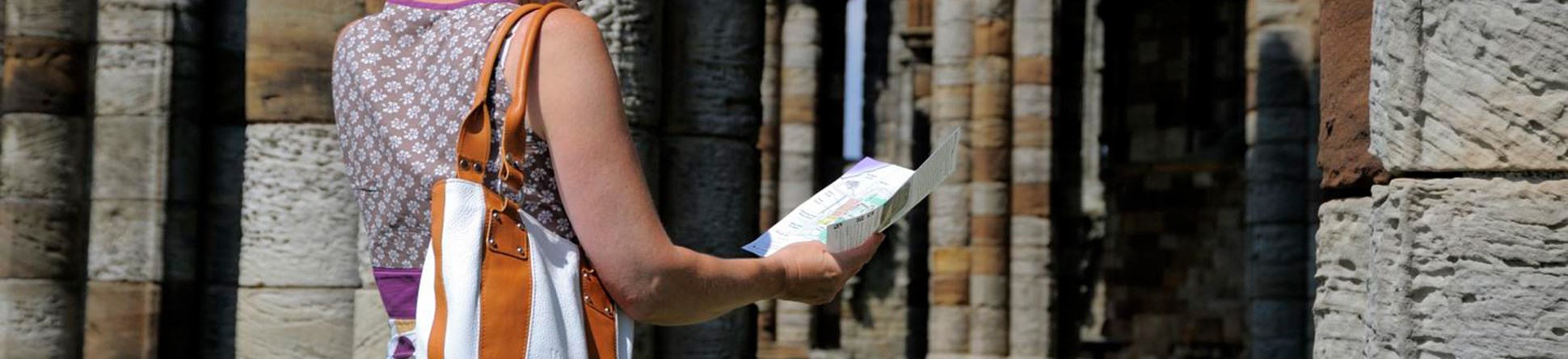 A person with short blonde hair, wearing a bag. Reading a brochure inside the columned archway of an ancient stone building