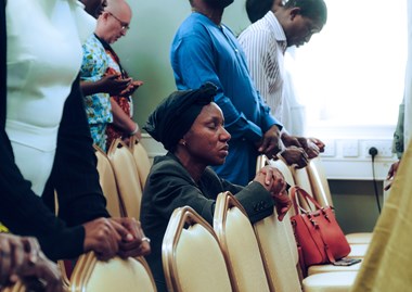 A woman in a headscarf kneels in prayer with eyes closed among rows of chairs.