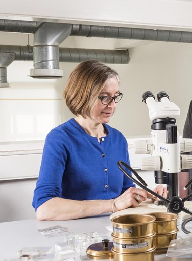 Photograph showing a woman in a lab featuring a microscope, bottles and boxes in the background. A woman with short hair and glasses is looking at a petri dish on a table.  