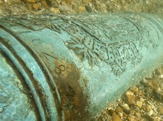 Green metallic cannon inscribed with patterns and text, sitting in a layer of gravel underwater.