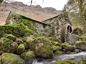 A stone-built water mill on a rocky stream in front of a tall hill.