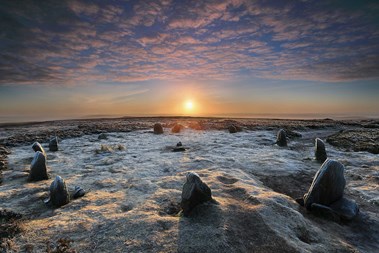 A photograph of a rural landscape at sunset with twelve stones positioned in a circle in the foreground.