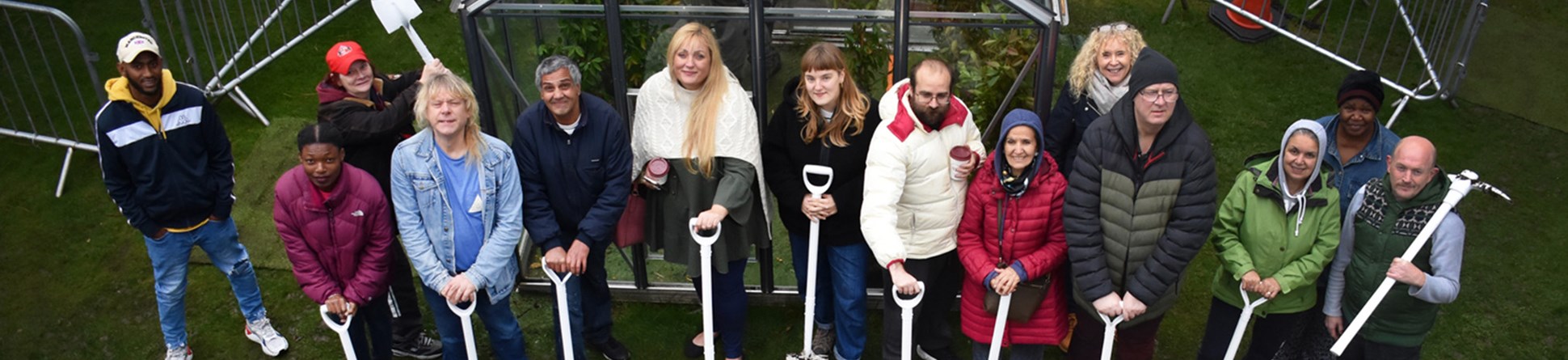 People stood in a community garden smiling, holding muddy white spades with white wheelbarrows in the foreground. 