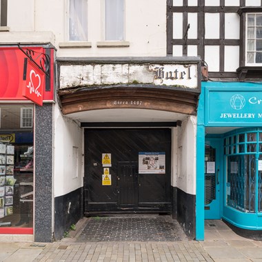 A cobbled, recessed arched entranceway into a historic inn building. A sign above reads 'circa 1497' and another, very damaged, says 'hotel'.