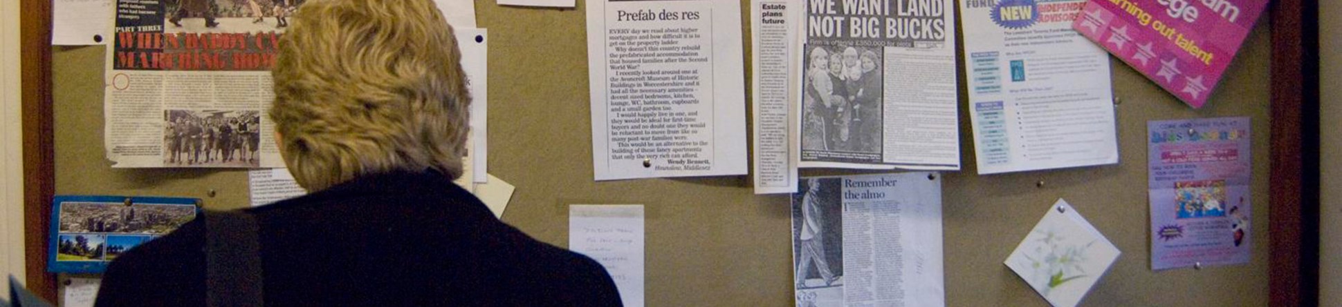 A photograph from behind of a person with short wavy hair looking at a General Information noticeboard with newspaper clippings and flyers
