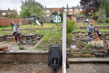 Either side of a garden fence, women cut their adolescent childrens' hair. On the left side, children play with a ball in the background.