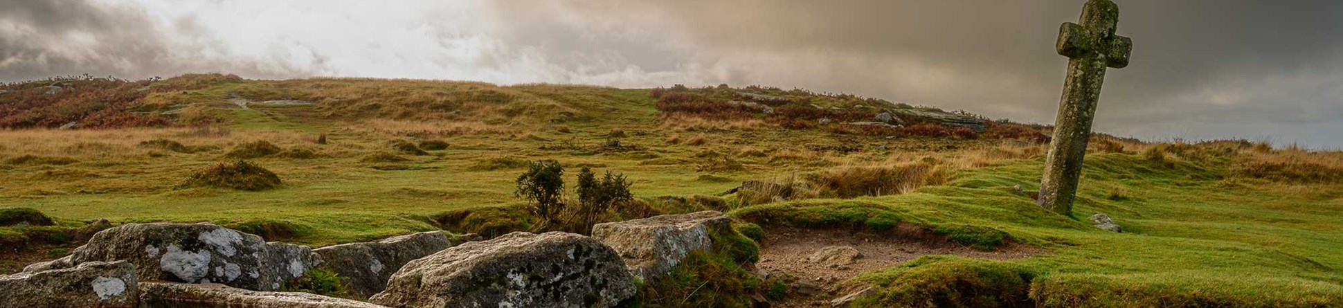 A photograph of a moorland landscape with a small waterfall flowing into a stream, and a vertical stone cross positioned alongside the water.